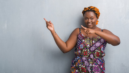 Poster - Young african american woman over grey grunge wall wearing orange braids smiling and looking at the camera pointing with two hands and fingers to the side.