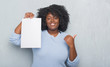 © Krakenimages.com - Young african american woman over grey grunge wall holding blank paper sheet pointing and showing with thumb up to the side with happy face smiling