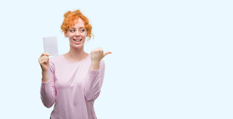 Poster - Young redhead woman holding blank notebook pointing and showing with thumb up to the side with happy face smiling