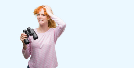 Poster - Young redhead woman looking through binoculars stressed with hand on head, shocked with shame and surprise face, angry and frustrated. Fear and upset for mistake.