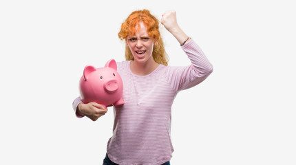 Canvas Print - Young redhead woman holding piggy bank annoyed and frustrated shouting with anger, crazy and yelling with raised hand, anger concept