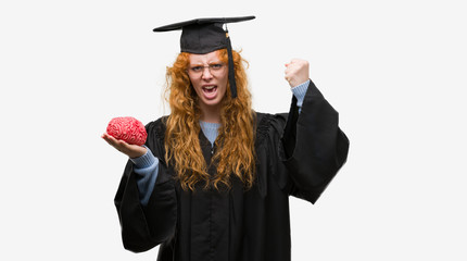 Canvas Print - Young redhead student woman wearing graduated uniform holding brain annoyed and frustrated shouting with anger, crazy and yelling with raised hand, anger concept