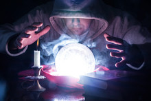 Wizard With Hood And Lights Smoke Magic Crystal Ball On Desk With Candle In Candlestick And Old Books Predicting The Future By Looking Into Ball And Holding Hand Above Tools. Close Up, Selective Focus