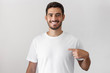 © Damir Khabirov - Handsome young man pointing with index finger at blank white t-shirt with empty space for your advertising text or image, standing isolated on gray background