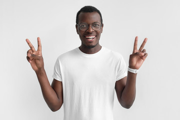 Wall Mural - Young african american man isolated on gray background in white t-shirt with optimistic smile, showing victory sign with both hands, looking friendly