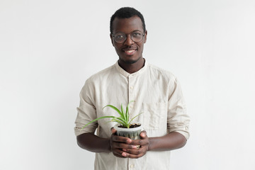 Studio shot of young smiling african man standing isolated on gray background, dressed in light shirt, holding flower pot with green plant in hands looking happy