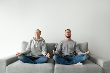 Wall Mural - Happy millennial couple sit in lotus position on home coach, calm boyfriend and girlfriend meditate practicing yoga on sofa, mindful man and woman relieve stress breathing fresh air with mudra hands