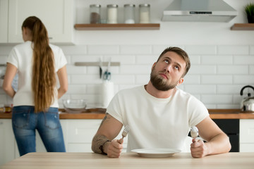 Wall Mural - Lazy millennial husband look bored waiting for dinner or breakfast cooked by wife, indifferent man wanting eat sitting with empty plate at table thinking about something rolling eyes looking bored