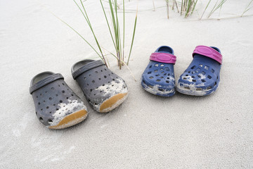 Two sand covered colorful rubber clog shoes on white beach sand with green blades of grass - concept leisure holiday vacation lonesome nature dune environment relaxation travel lifestyle fashion