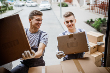 Wall Mural - Two young handsome smiling movers wearing uniforms are unloading the van full of boxes. House move, mover service.