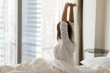 Woman Waking Up Happy Stretching Sitting On Comfortable Bed Looking Out Of Big Skyscraper Window In Modern Hotel Bedroom Enjoying Good Morning And City View Starting New Day, Wellbeing Concept