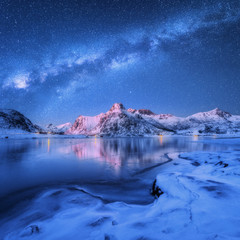 Milky Way above frozen sea coast and snow covered mountains in winter at night in Lofoten Islands, Norway. Arctic landscape with blue starry sky,  water, ice, snowy rocks, milky way. Beautiful space