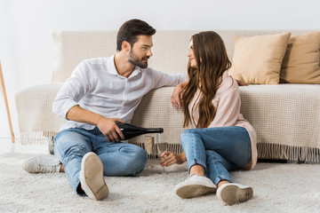 Wall Mural - man pouring champagne into glass while sitting on floor together with girlfriend at home