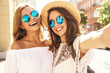 Two young female smiling hippie brunette and blond women models in summer white hipster dress taking selfie photos for social media on smartphone on the street background. Surprise face, emotions,