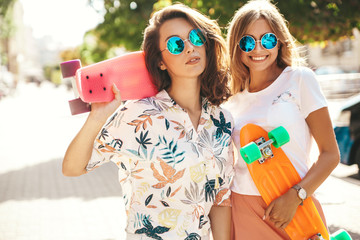 Two young stylish smiling hippie brunette and blond women models in summer sunny day in hipster clothes with penny skateboard posing on the street background