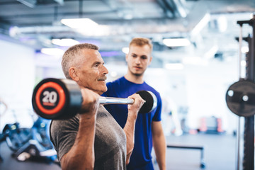 Older man lifting weights, supervised by gym assistant.