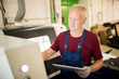 © pressmaster - Mature factory engineer in overalls holding tablet while standing in front of industrial computer and touching its display