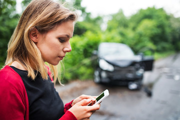 Wall Mural - A young woman with smartphone by the damaged car after a car accident, text messaging.