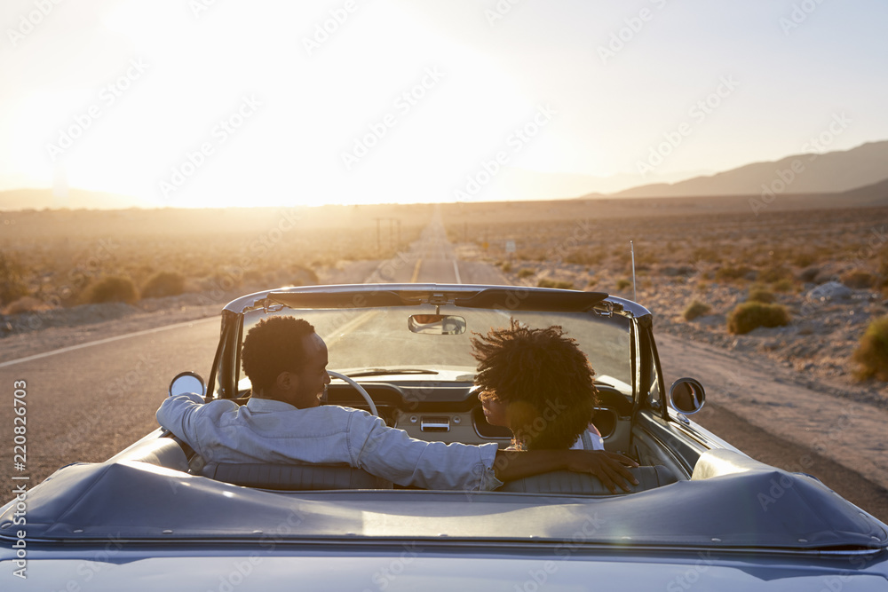Rear View Of Couple On Road Trip Driving Classic Convertible Car Towards Sunset - obrazy, fototapety, plakaty 
