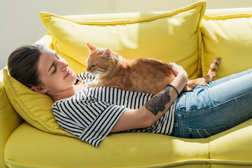 beautiful smiling young woman lying on yellow couch with cute red cat