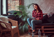 © Fxquadro - Smiling brunette girl student dressed in a plaid shirt and jeans holding digital tablet sitting on a couch in a room with loft interior, looking out the window.