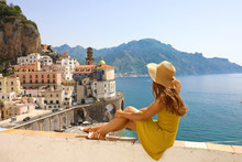 Beautiful Young Woman With Hat Sitting On Wall Looking At Stunning Panoramic Village Of Atrani On Amalfi Coast, Italy