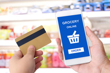 Hand holding credit card and smartphone with grocery online shopping application on device screen over blur supermarket background, business finance  and technology, online payment concept