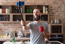 Vlogger Streaming Live Using Mobile Phone Camera On Selfie Stick. Bearded Hipster Man Communicating With Subscribers Standing At Home Kitchen.