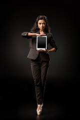 Wall Mural - attractive businesswoman in suit and ballet shoes showing tablet with blank screen on black backdrop