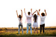 © Leika production - Back view. Two guys and two girls are standing in the field on a summer day and holding their hands up.