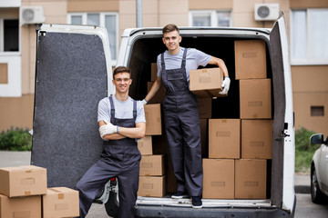 Wall Mural - Two young handsome smiling movers wearing uniforms are unloading the van full of boxes. House move, mover service.