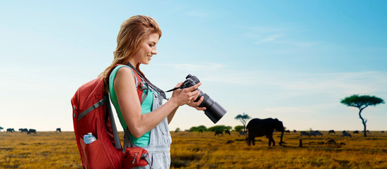 Wall Mural - travel, tourism and photography concept - happy young woman with backpack and camera photographing over animals in african savannah background