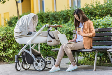 Wall Mural - freelancer working with laptop on bench near baby stroller in park and holding coffee in paper cup