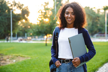 Poster - Beautiful african woman student walking in the park holding laptop computer.