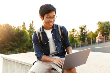 Canvas Print - Smiling asian man student with backpack using laptop