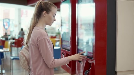 Young female visitor is making order in machine in hall of cafe with fast food. She is choosing meal on big display