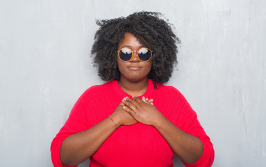 Poster - Young african american plus size woman over grey grunge wall wearing retro sunglasses smiling with hands on chest with closed eyes and grateful gesture on face. Health concept.