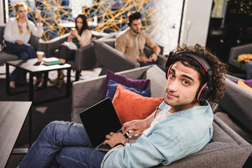 Wall Mural - young businessman in headphones sitting on couch and using laptop with blank screen at modern coworking office