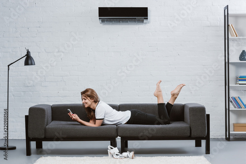 Cuadros en Lienzo happy young woman using smartphone while lying on sofa under air conditioner han