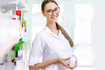 Wall Mural - A smiling charming slim dark-haired lady with glasses, wearing a white coat, stands next to the shelf and shows a small jar in the modern pharmacy. Her slim profile is shown.