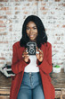 © sofiko14 - Young afro american girl in red coat in urban background holds the retro camera in her hands