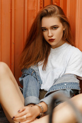 Canvas Print - Beautiful young female model in a jeans jacket sits near an orange metal wall outdoors