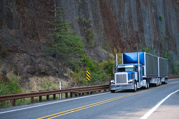 Big rig blue classic American bonnet semi truck with two covered black dry van semi trailers running on mountain road around huge cliff