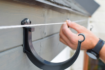 Man hands installing dark grey plastic roof gutter holder on wooden eaves and check level for drainage system of roof. Home improvement concept.