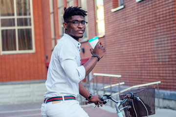 stylish and young African American man holding a glass with coffee sitting on a bike in the summer park