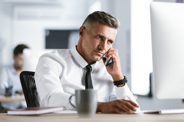 Fototapete - Image of adult businessman 30s wearing white shirt and tie sitting at desk in office by computer, and talking on smartphone
