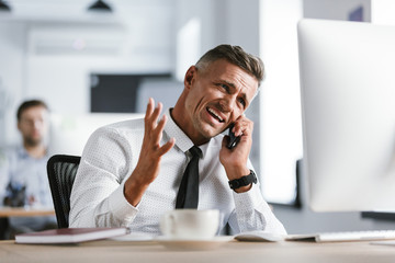 Fototapete - Image of irritated businessman 30s wearing white shirt and tie sitting at desk in office by computer, and screaming while talking on smartphone