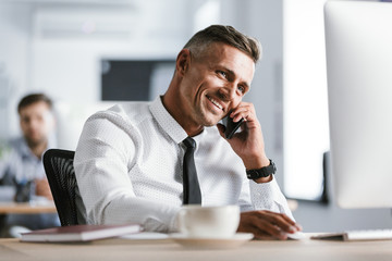 Fototapete - Image of european adult man 30s wearing white shirt and tie sitting at desk in office by computer, and speaking on smartphone
