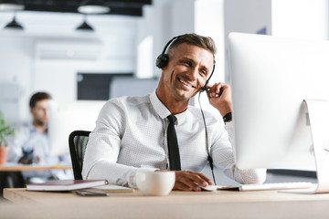 Fototapete - Photo of smiling operator man 30s wearing office clothes and headset, sitting by computer in call center