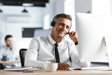 Fototapete - Photo of joyful operator man 30s wearing office clothes and headset, smiling while working on computer in call center